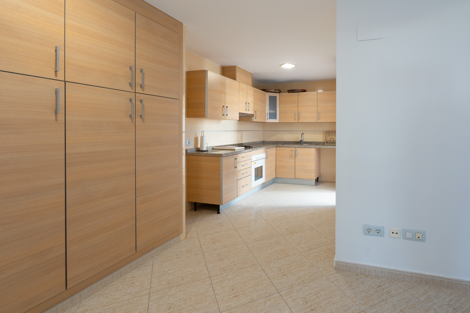 Amplia cocina con armarios de madera clara, encimeras de granito moteado y horno integrado. Un gran mueble despensero a juego se sitúa a la izquierda sobre el suelo de baldosas beige.