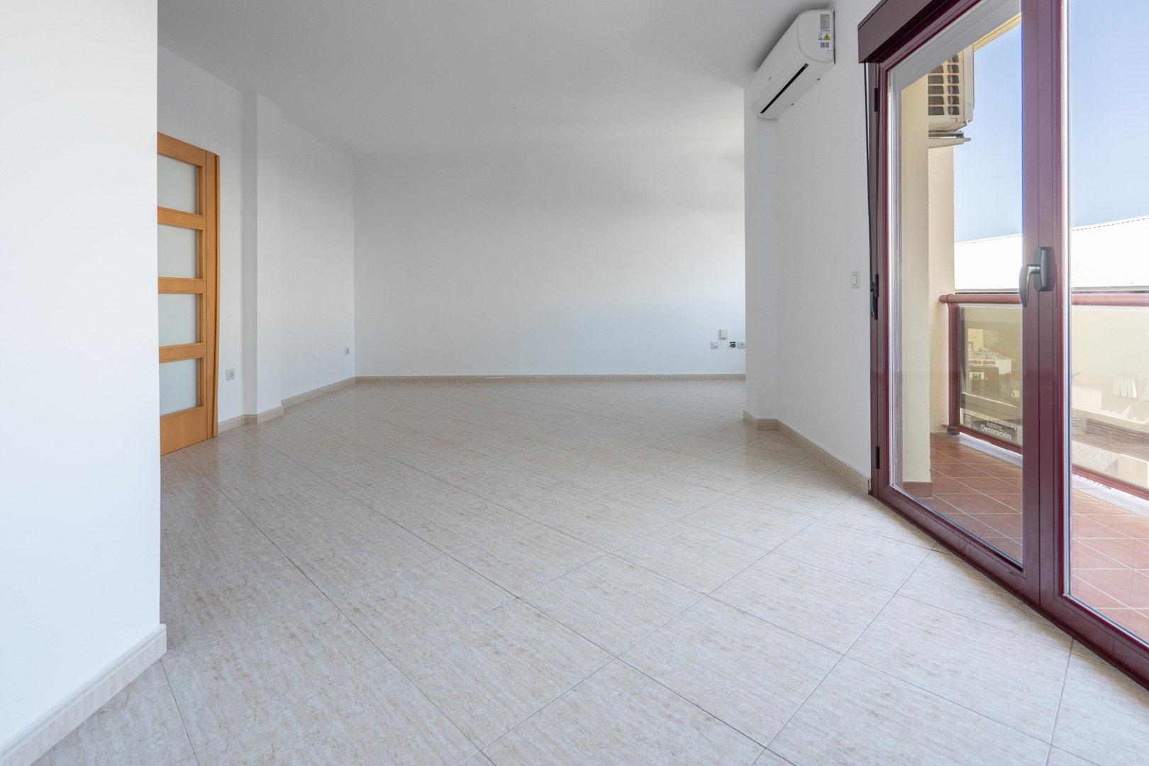 Amplio salón-comedor vacío con suelo de gres claro, puerta de madera con paneles de cristal y acceso a terraza mediante puerta corredera de aluminio granate. Incluye unidad de aire acondicionado.
