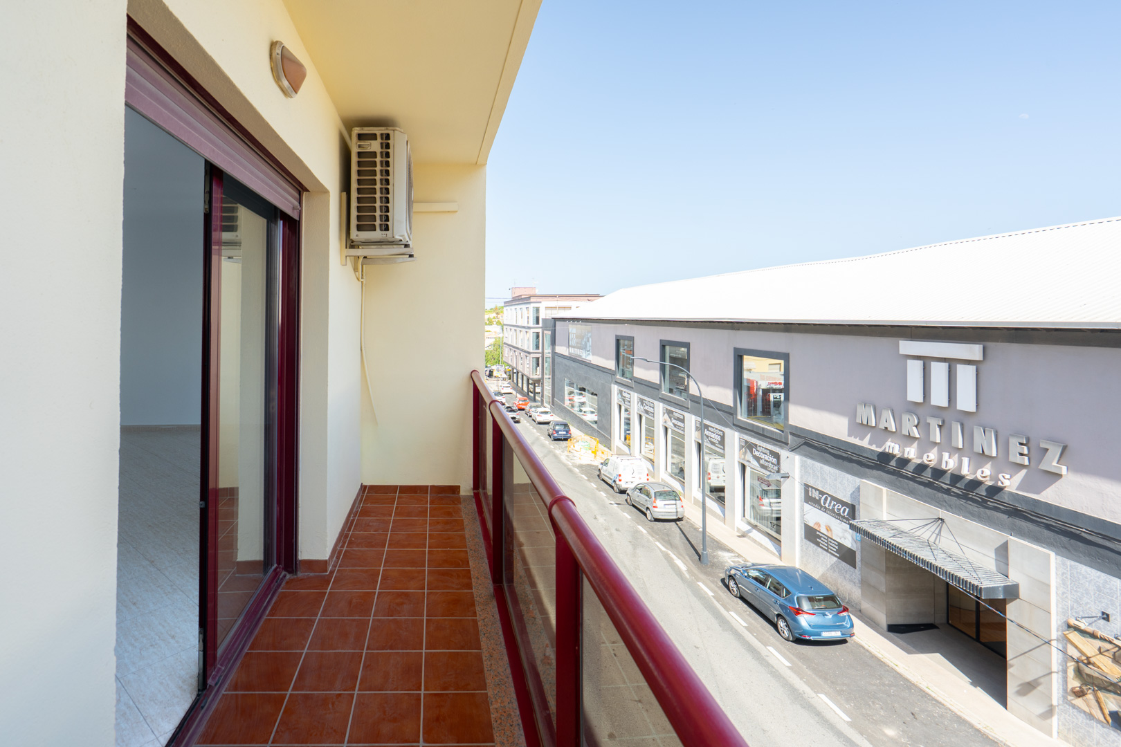 Balcón exterior con suelo de baldosas de terracota y barandilla roja. Incluye unidad de aire acondicionado y vistas a la calle principal con comercios como "Martinez Muebles".