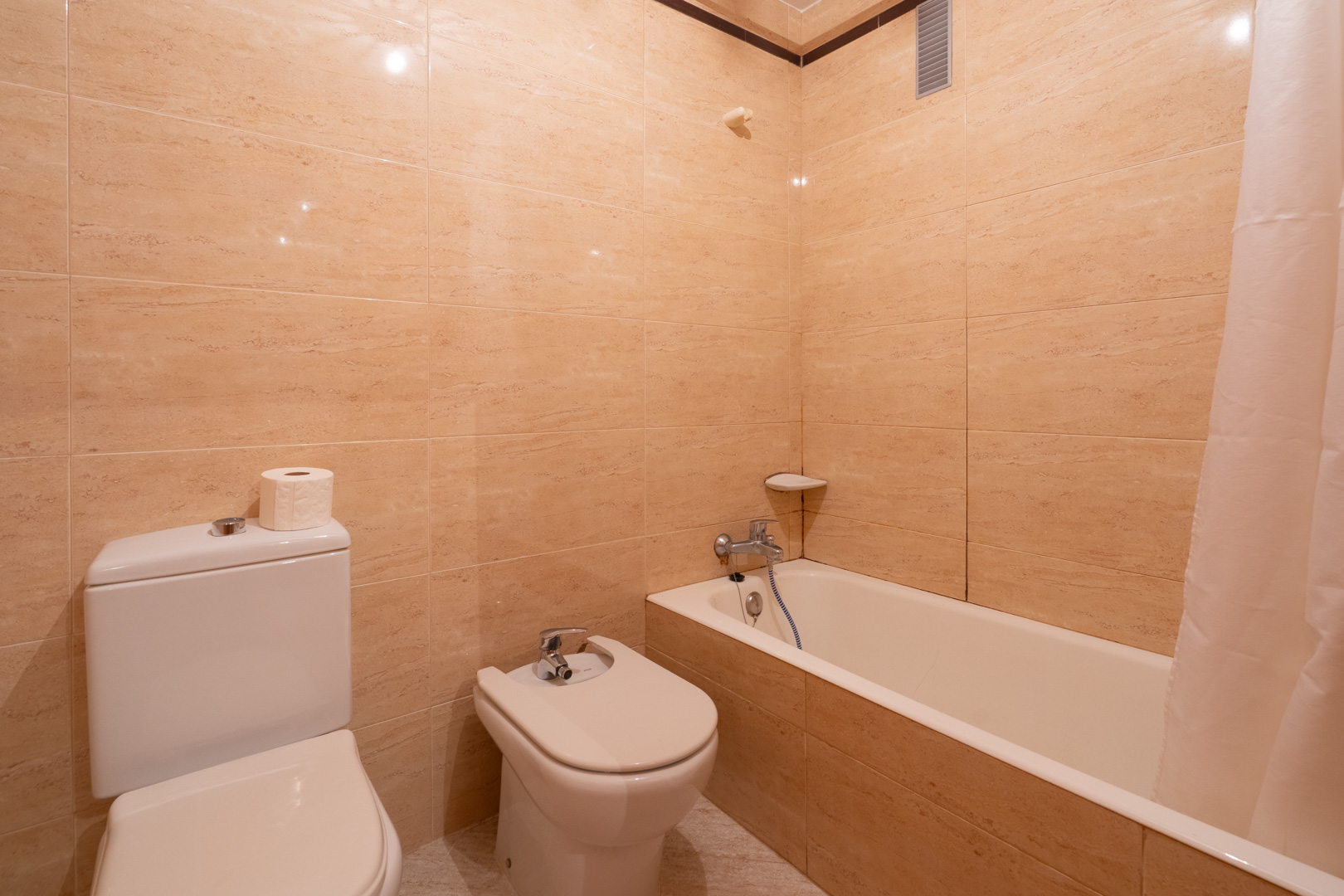 Baño con paredes de azulejos beige, equipado con inodoro, bidé y bañera con cortina. Incluye grifería cromada y estante esquinero.