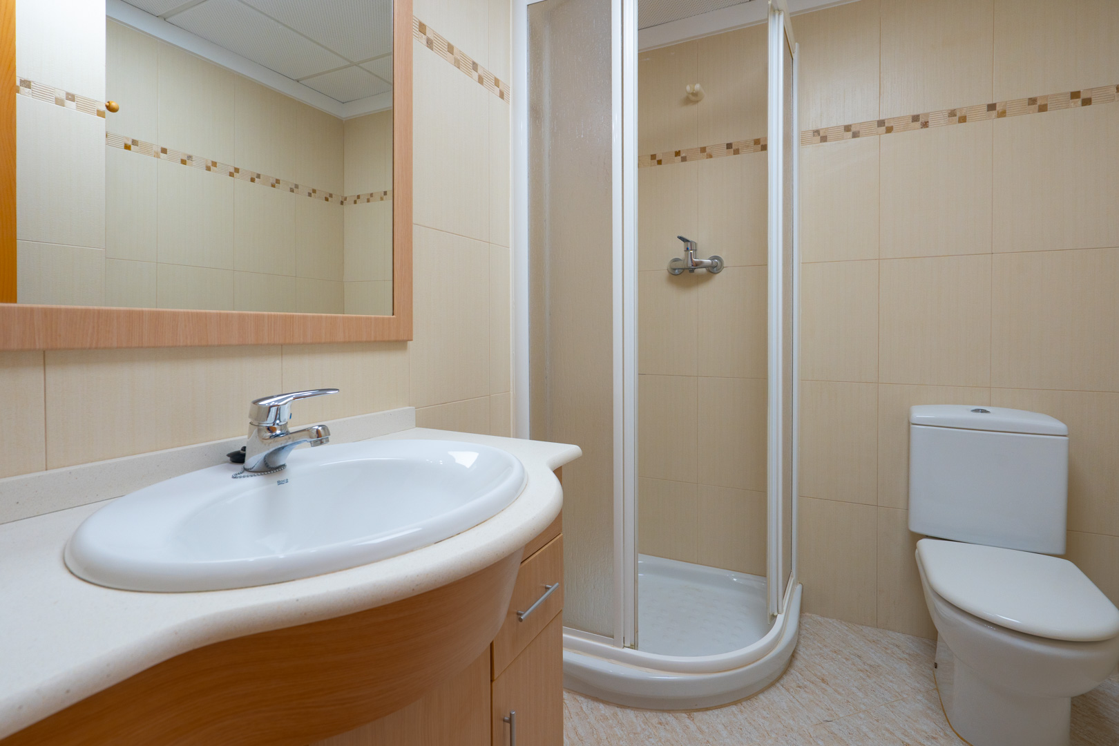 Baño funcional con lavabo ovalado sobre mueble de madera clara, espejo enmarcado, ducha de esquina y azulejos beige con cenefa decorativa.