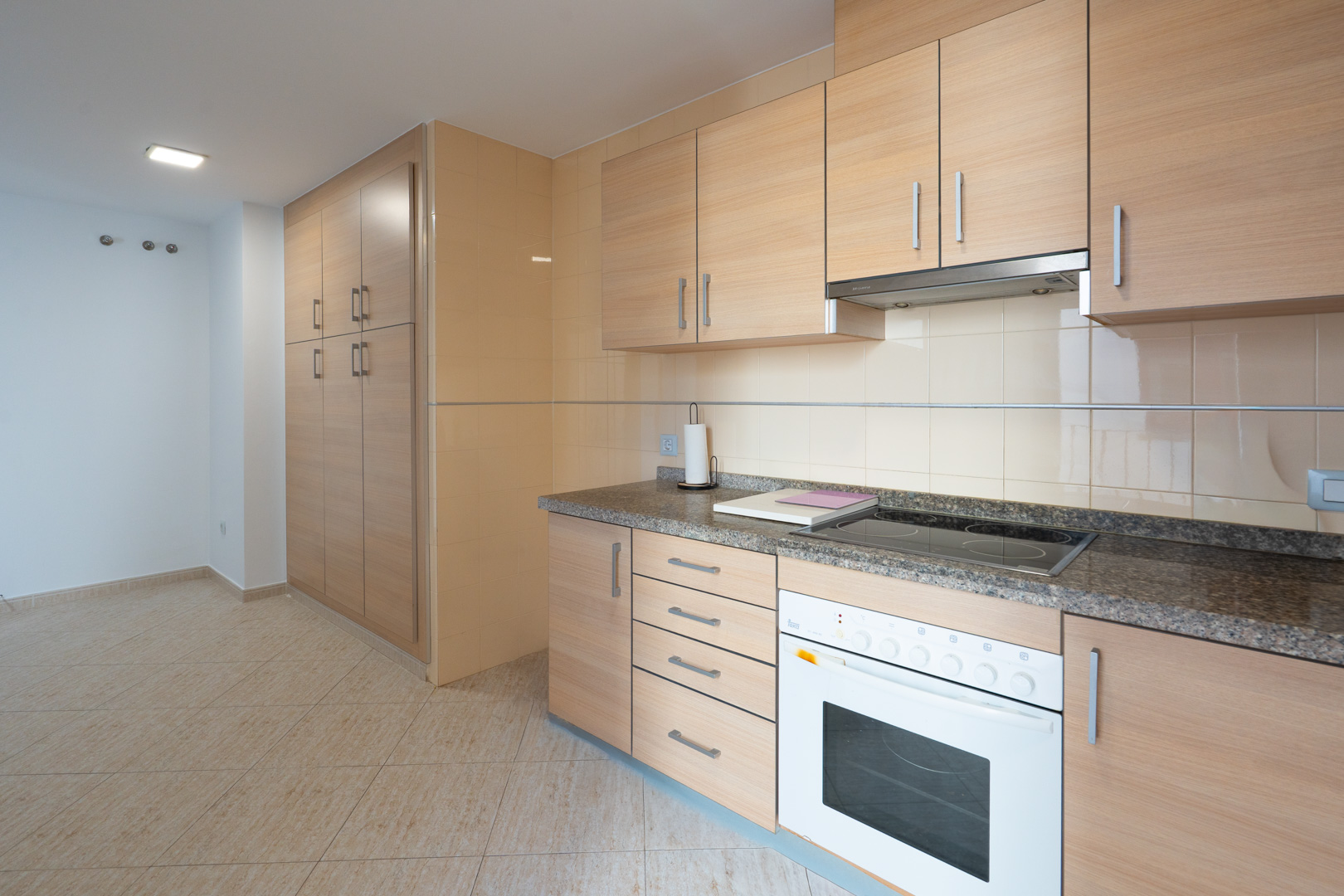 Cocina funcional con armarios de madera clara, encimera de granito, placa vitrocerámica y horno integrados. Incluye una despensa alta y suelo de gres.