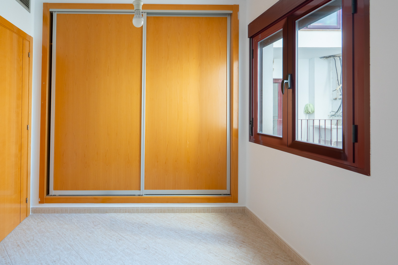 Dormitorio secundario con amplio armario empotrado de puertas correderas de madera clara y ventana de madera oscura con vistas a un patio interior.