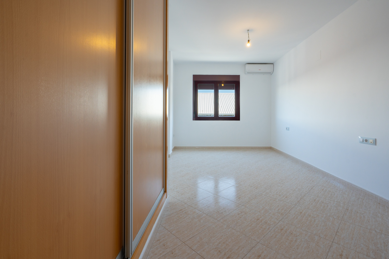 Dormitorio vacío con amplio armario empotrado de madera clara, suelo de baldosas beige en diagonal y ventana con vistas a tejados. Incluye unidad de aire acondicionado.