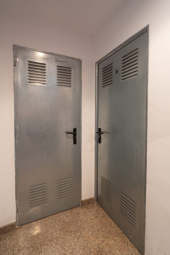 Dos puertas metálicas grises con ventilación superior e inferior, una marcada con 'Benissa Denia' y la otra con el número '5', sobre un suelo de granito moteado.
