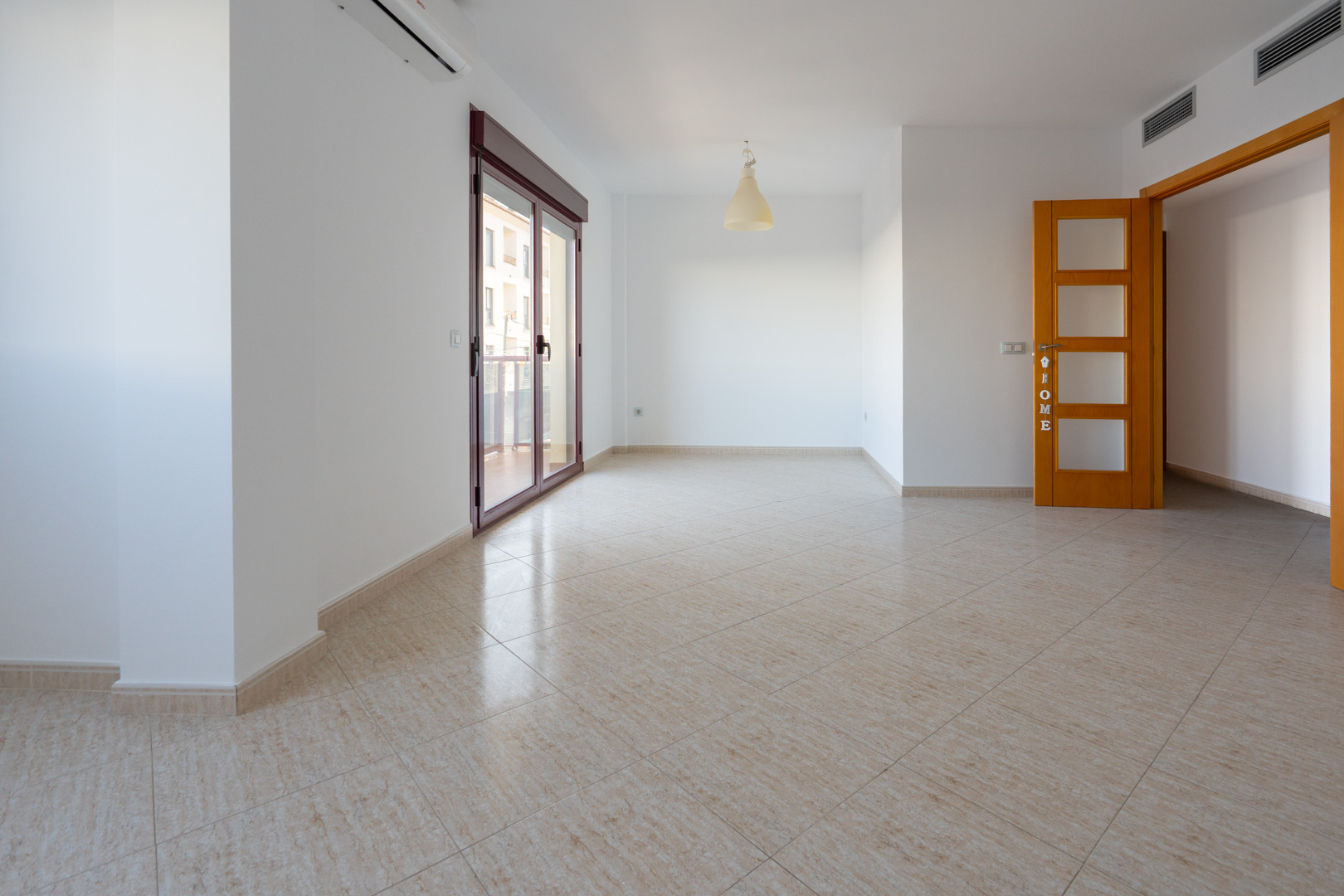 Espaciosa estancia con suelo de gres cerámico beige. Cuenta con una amplia puerta corredera granate que da a la terraza, aire acondicionado split y una puerta de madera clara con paneles de cristal.