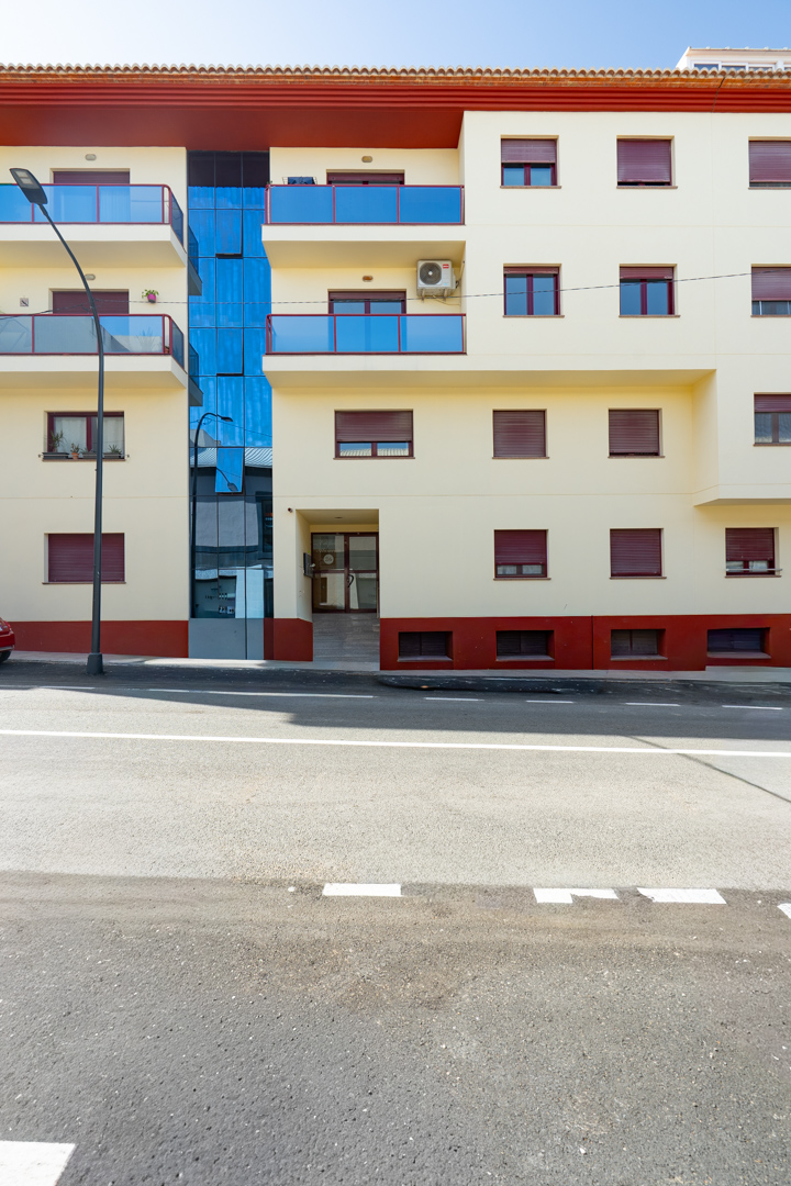 Fachada exterior del edificio residencial con balcones de cristal azul, persianas rojas y base en tonos terracota. El diseño incluye un elemento central acristalado y una entrada accesible desde la calle.
