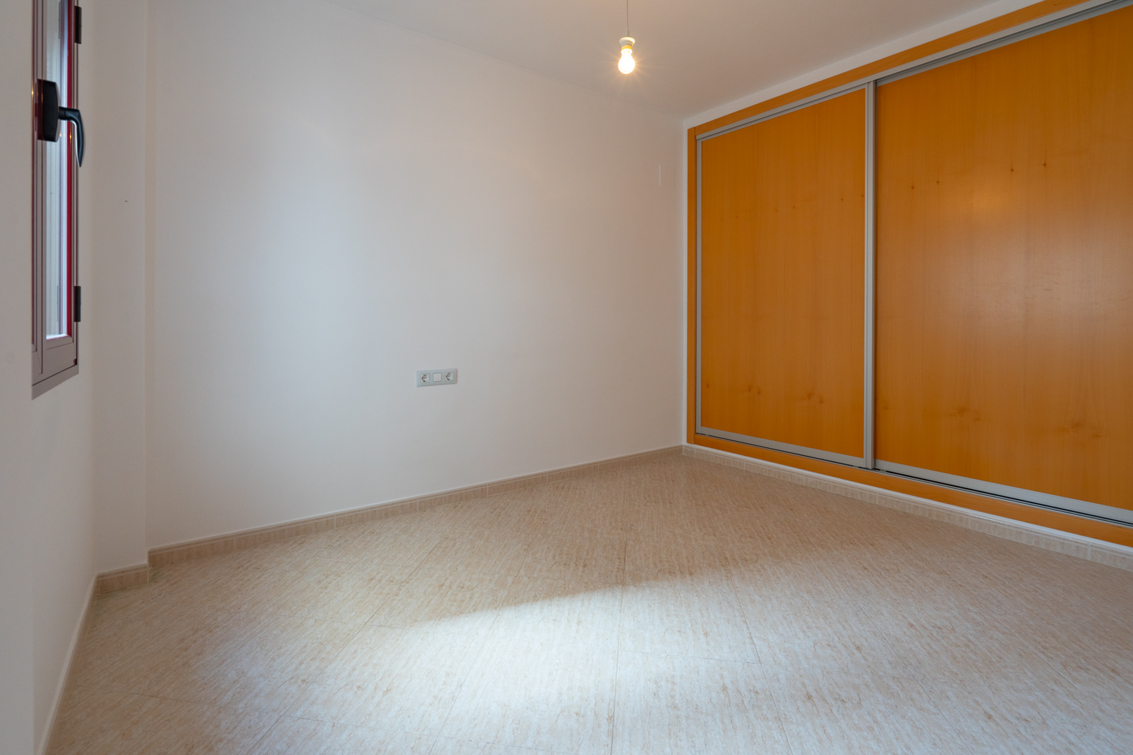 Habitación luminosa con suelo de gres beige y paredes blancas. Incluye un armario empotrado de dos puertas correderas de madera clara y una ventana con marco oscuro que aporta luz natural.
