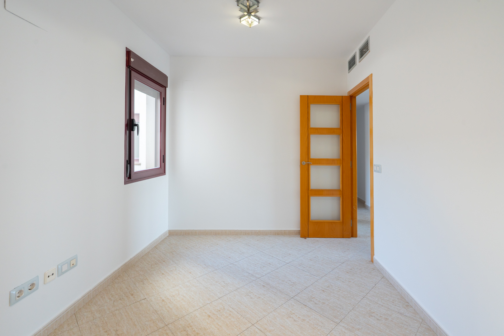 Habitación vacía con paredes blancas, ventana de marco burdeos y suelo de baldosas beige claro. Incluye puerta de madera con paneles de cristal y rejillas de aire acondicionado.