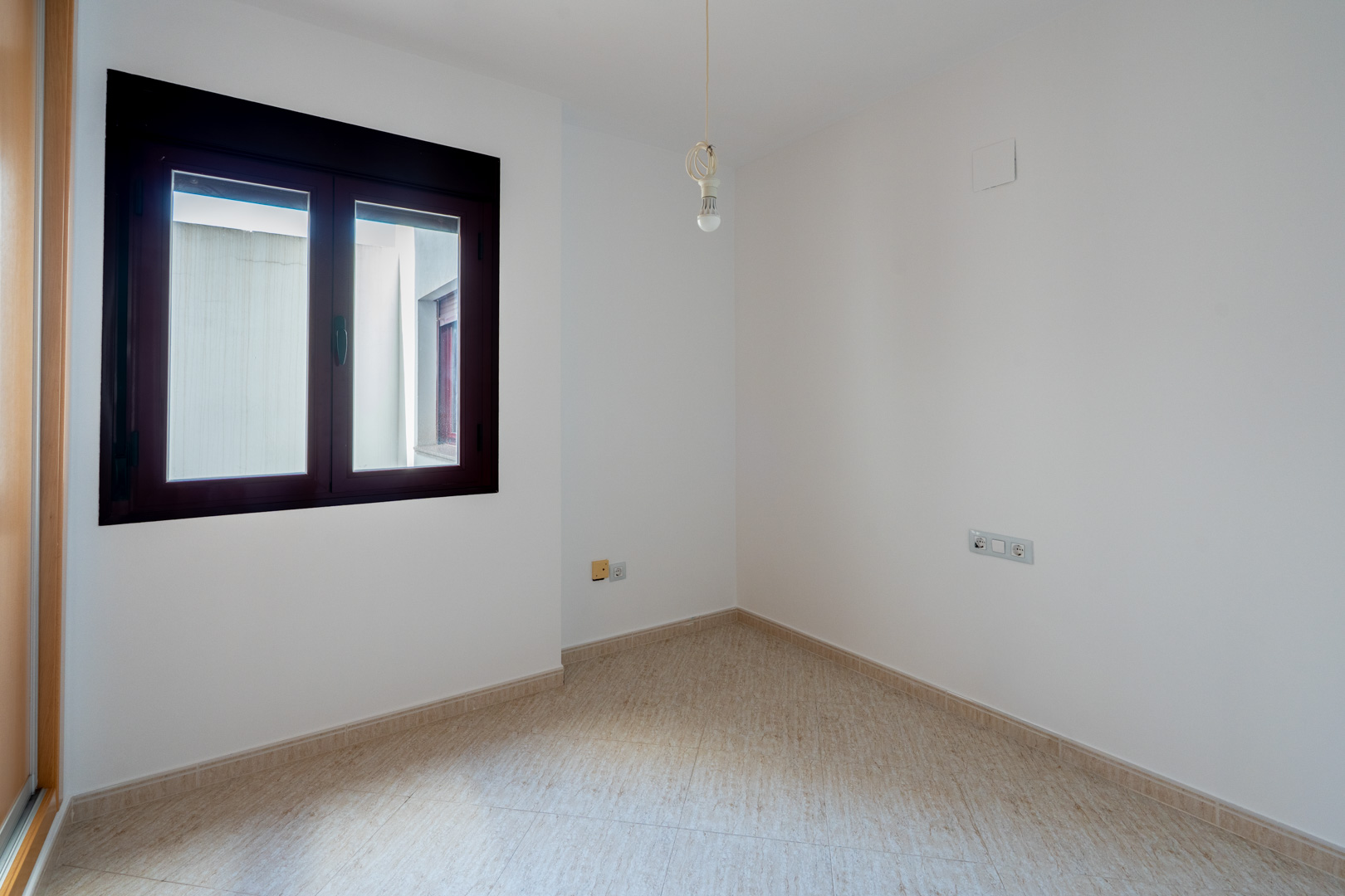 Habitación vacía con paredes blancas y suelo de baldosas claras. Presenta una ventana de marco oscuro que da a un patio interior y un punto de luz central en el techo.