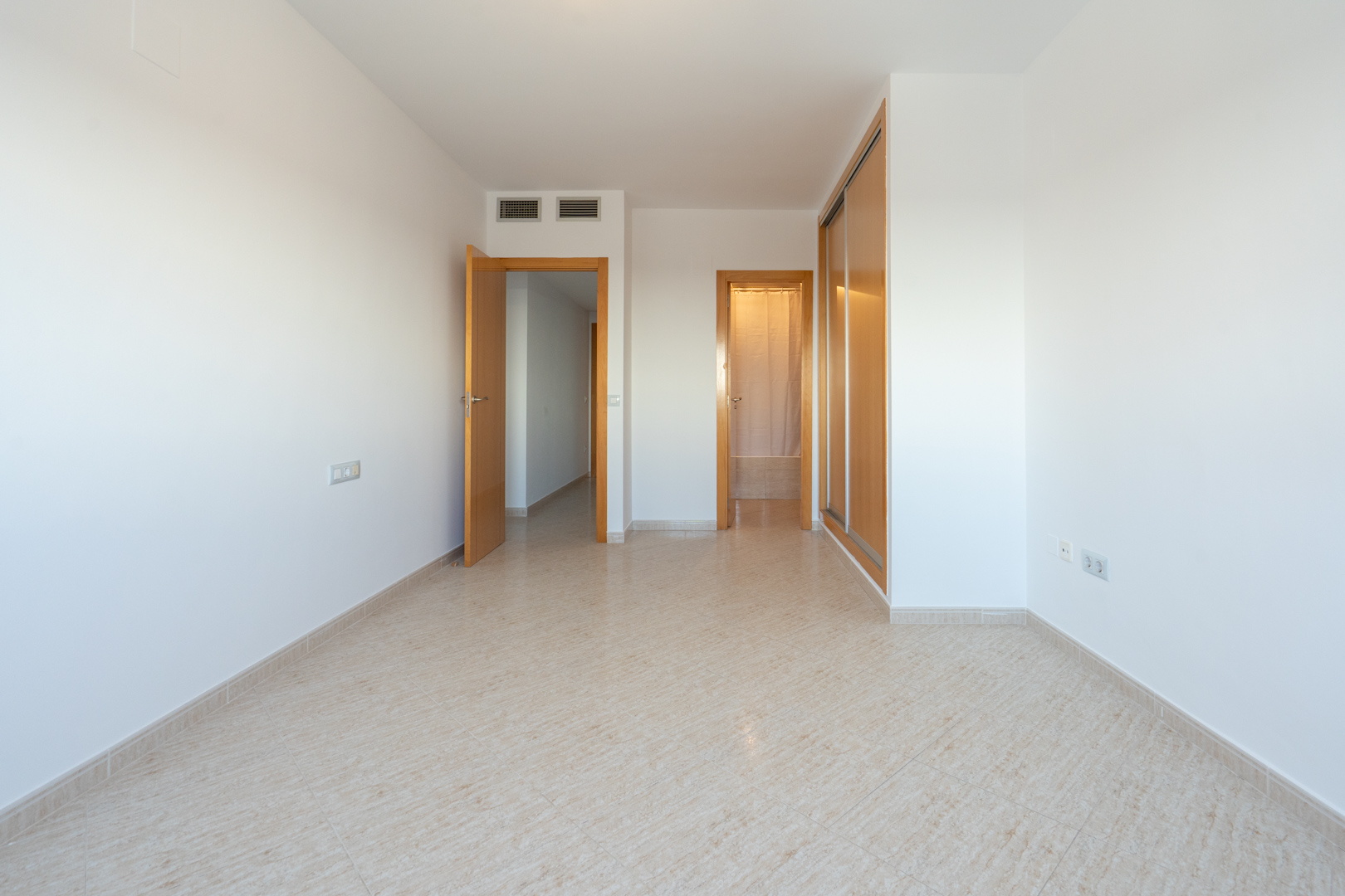 Habitación vacía con suelo de baldosas claras, armario empotrado de madera y puertas que conectan a otras estancias. Se aprecian rejillas de aire acondicionado.