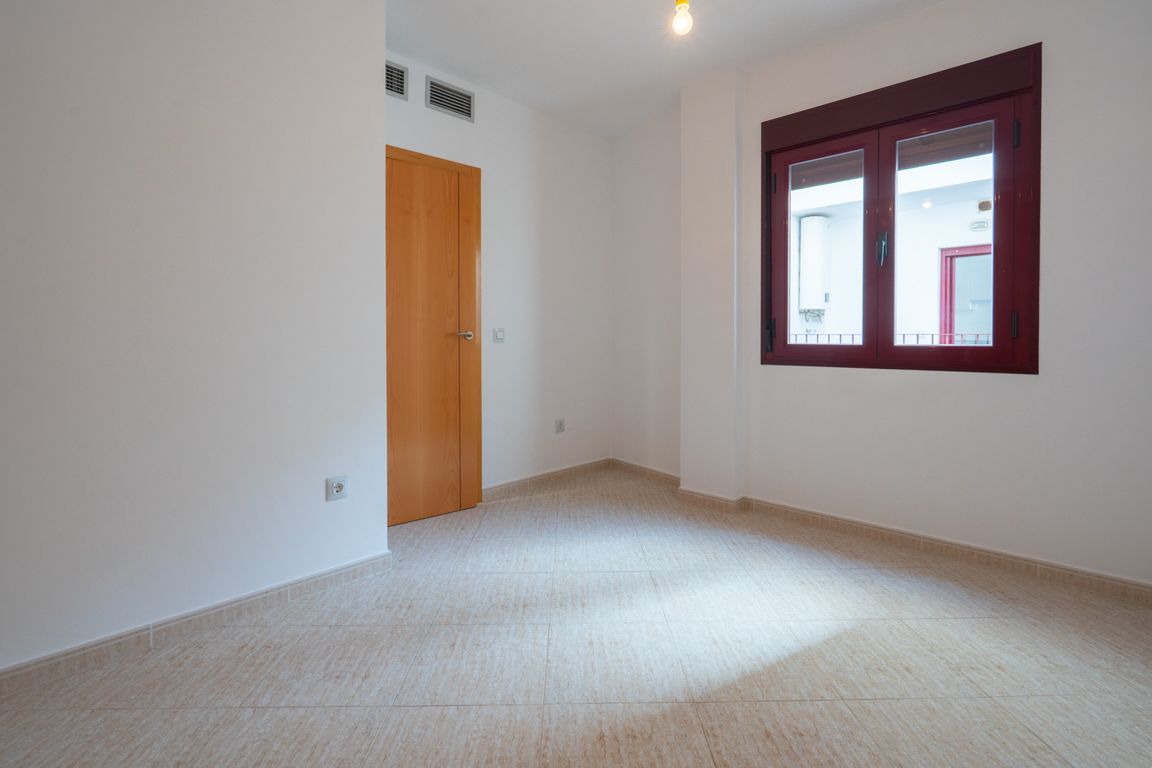 Habitación vacía con suelo de baldosas claras y paredes blancas. Dispone de una puerta de madera y una ventana con marcos burdeos, con vistas a un patio interior con termo. Rejillas de aire visibles.