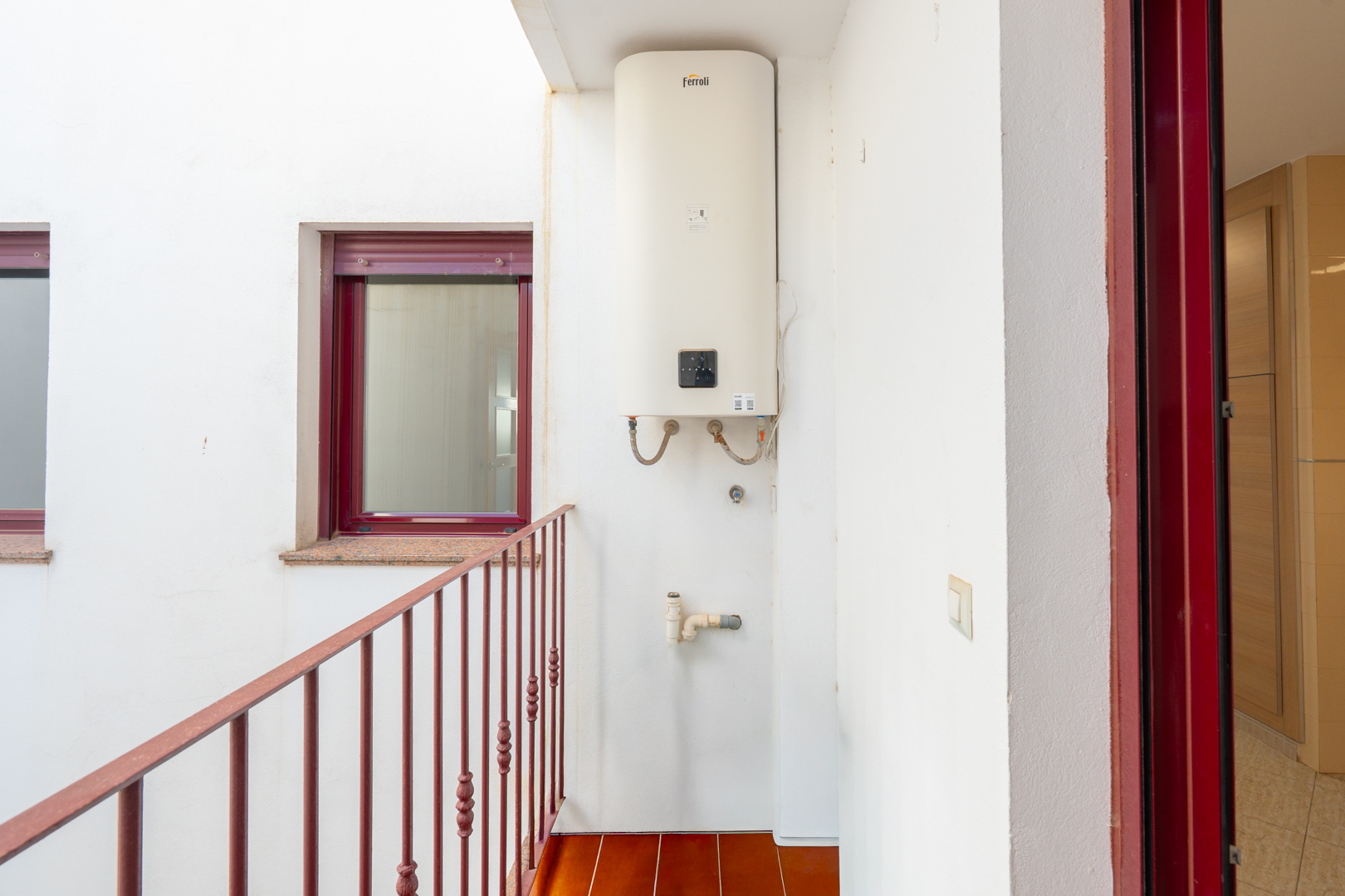 Patio interior con termo Ferroli montado en pared, suelo de baldosas terracota y barandilla metálica roja. Incluye ventana con marco burdeos y alféizar de granito, además de una puerta con marco rojo.