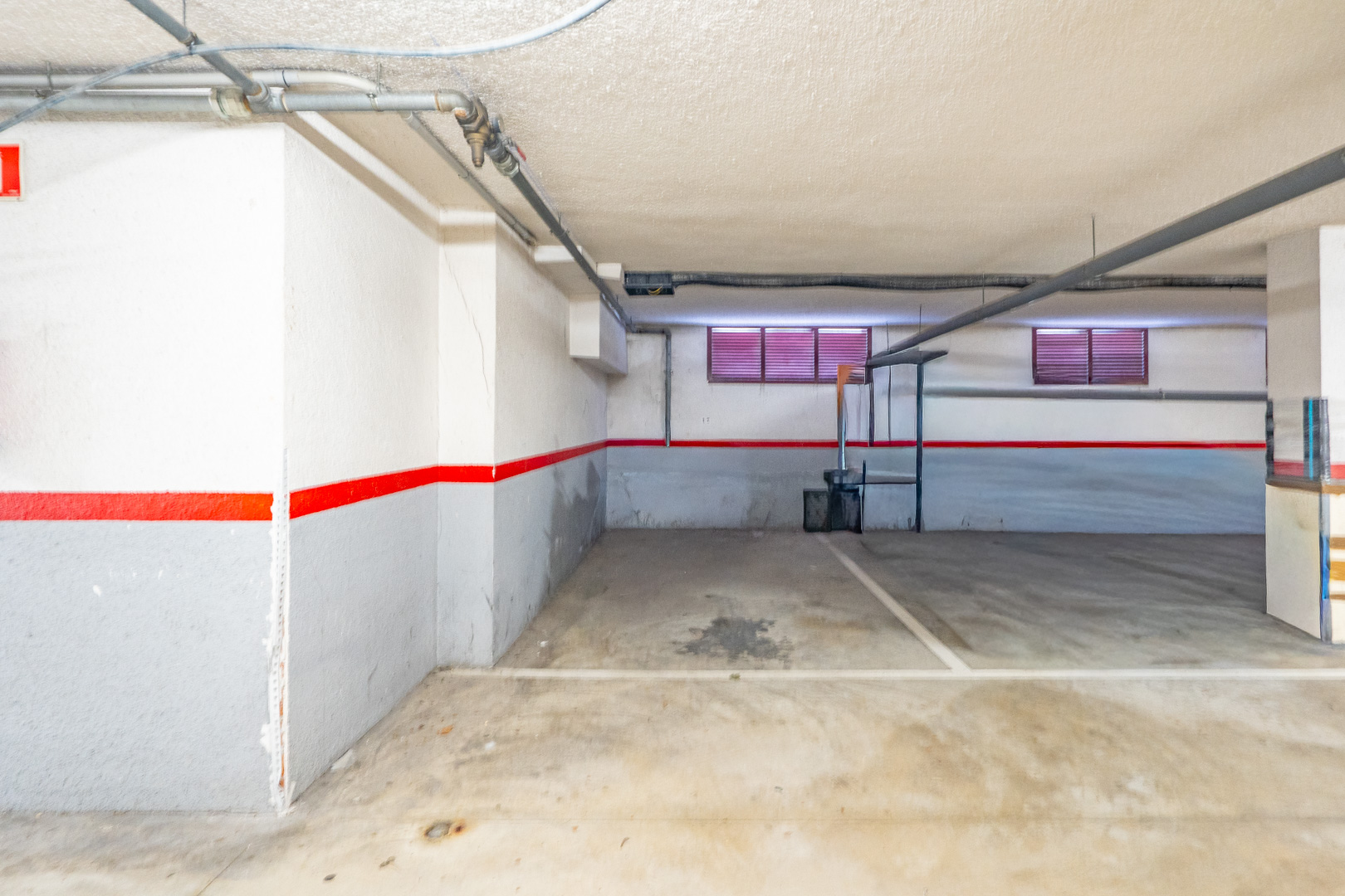 Plaza de garaje subterráneo con suelo de hormigón, paredes blancas y grises con franja roja, y tuberías vistas en el techo. Se aprecian dos pequeñas ventanas con persianas rojas.