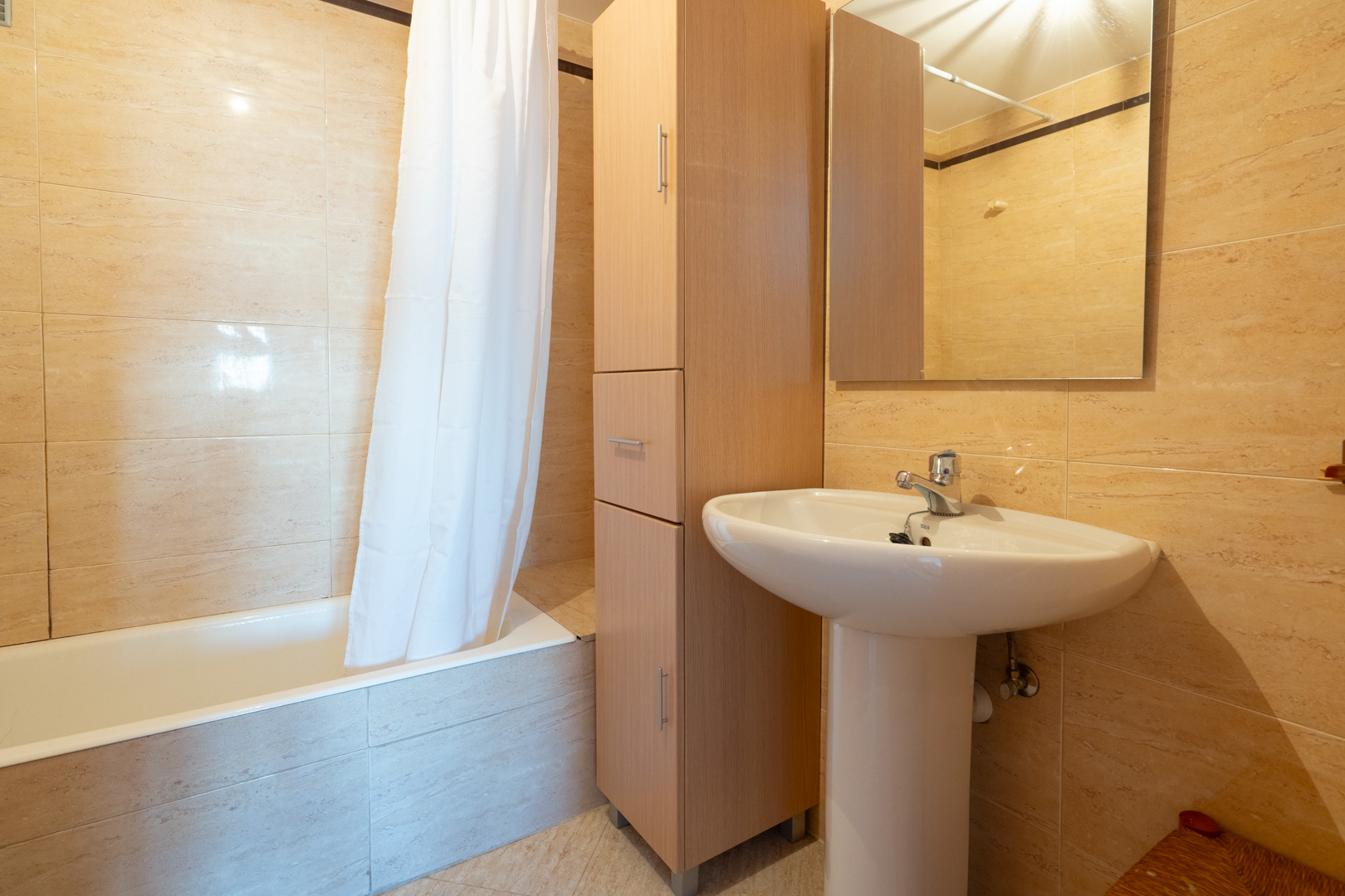 Segundo baño con lavabo de pedestal blanco, mueble alto de madera para almacenaje y bañera con cortina. Las paredes están revestidas con azulejos beige claro.
