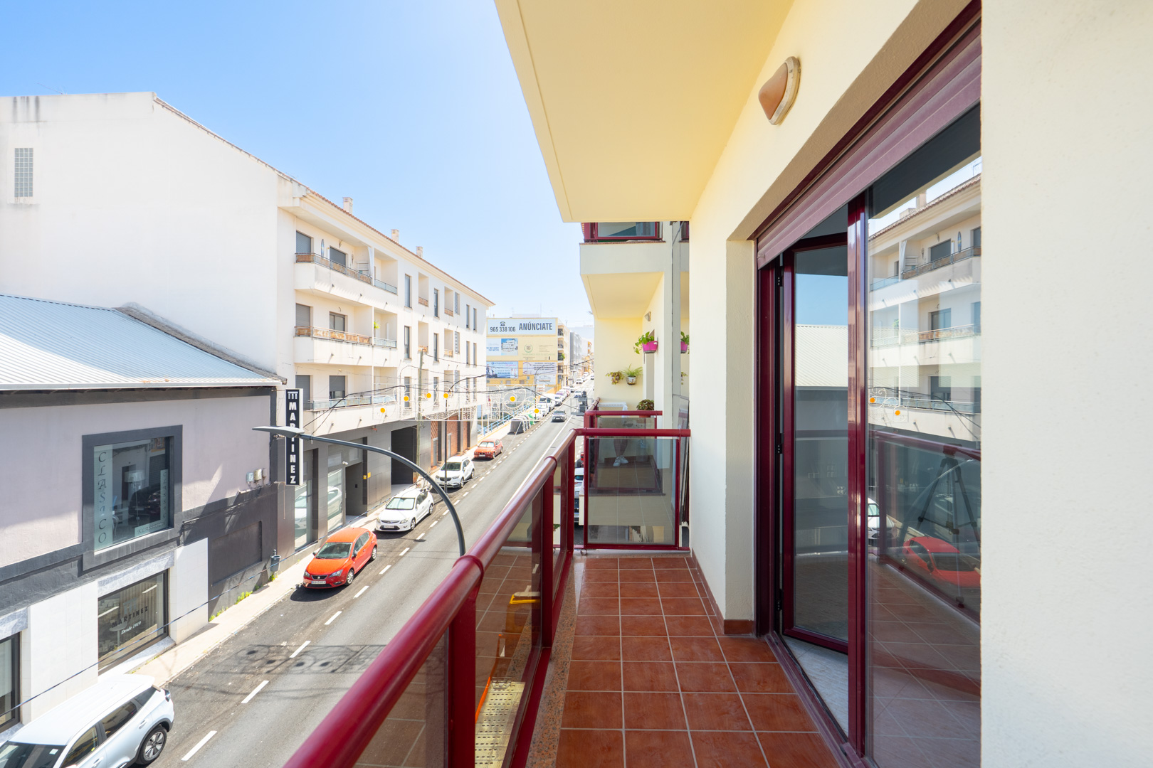 Terraza soleada con suelo de baldosas de terracota y barandilla de cristal con marcos rojos, ofreciendo vistas a una calle urbana con edificios.