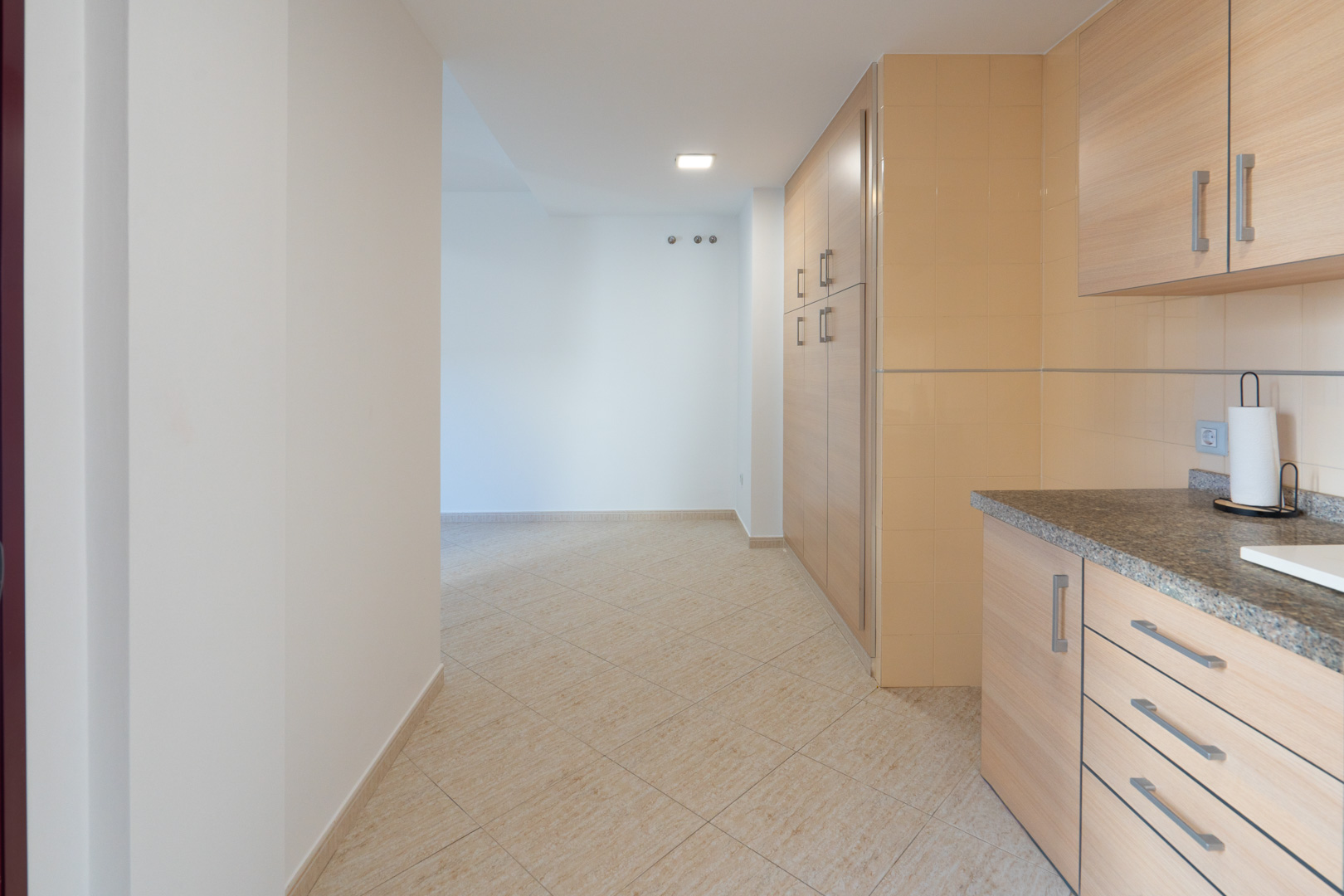 Transición desde la cocina con encimera de granito y armarios de madera clara hacia un pasillo con suelo cerámico beige y ganchos de pared.