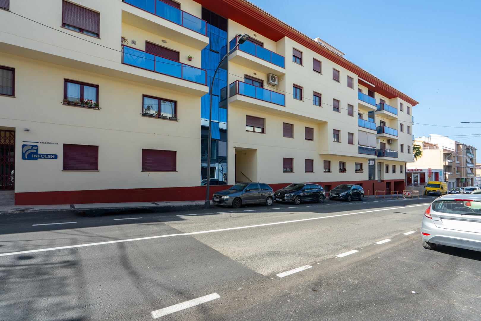 Vista exterior del edificio residencial con fachada clara, balcones de cristal azul, ventanas con persianas granates y un panel de vidrio vertical.