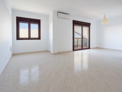 Amplia estancia con suelo de gres claro, paredes blancas, ventana y puerta corredera de acceso a terraza con marcos marrones, y aire acondicionado.