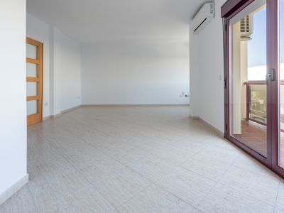Amplio salón-comedor vacío con suelo de gres claro, puerta de madera con paneles de cristal y acceso a terraza mediante puerta corredera de aluminio granate. Incluye unidad de aire acondicionado.