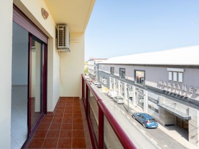 Balcón exterior con suelo de baldosas de terracota y barandilla roja. Incluye unidad de aire acondicionado y vistas a la calle principal con comercios como "Martinez Muebles".