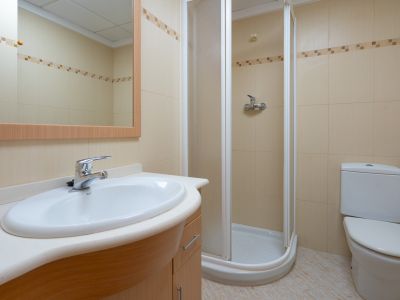 Baño funcional con lavabo ovalado sobre mueble de madera clara, espejo enmarcado, ducha de esquina y azulejos beige con cenefa decorativa.