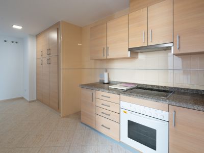Cocina funcional con armarios de madera clara, encimera de granito, placa vitrocerámica y horno integrados. Incluye una despensa alta y suelo de gres.