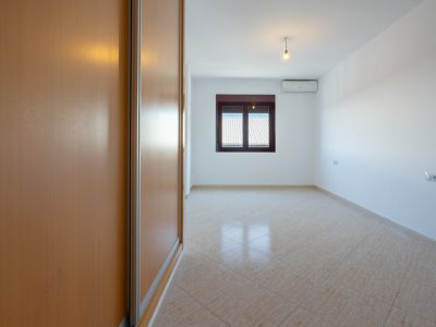 Dormitorio vacío con amplio armario empotrado de madera clara, suelo de baldosas beige en diagonal y ventana con vistas a tejados. Incluye unidad de aire acondicionado.
