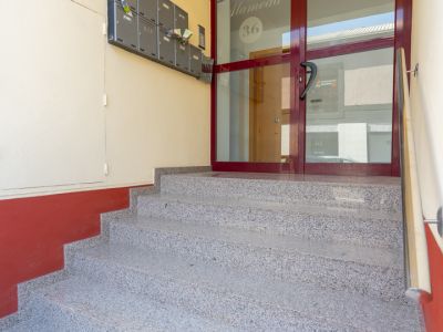 Escalera de efecto granito que conduce a la entrada principal del edificio, con una puerta de cristal enmarcada en rojo oscuro con la inscripción 'Edificio Alameda 36' y buzones grises a la izquierda.