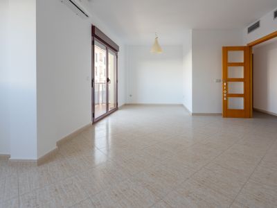 Espaciosa estancia con suelo de gres cerámico beige. Cuenta con una amplia puerta corredera granate que da a la terraza, aire acondicionado split y una puerta de madera clara con paneles de cristal.