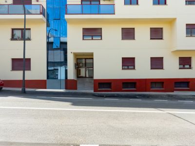 Fachada exterior del edificio residencial con balcones de cristal azul, persianas rojas y base en tonos terracota. El diseño incluye un elemento central acristalado y una entrada accesible desde la calle.