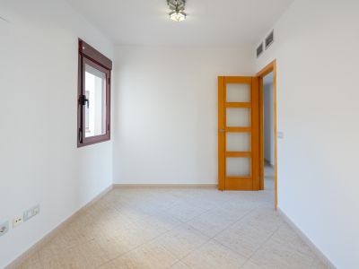 Habitación vacía con paredes blancas, ventana de marco burdeos y suelo de baldosas beige claro. Incluye puerta de madera con paneles de cristal y rejillas de aire acondicionado.