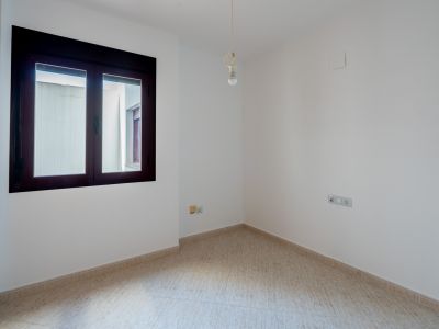Habitación vacía con paredes blancas y suelo de baldosas claras. Presenta una ventana de marco oscuro que da a un patio interior y un punto de luz central en el techo.