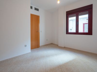 Habitación vacía con suelo de baldosas claras y paredes blancas. Dispone de una puerta de madera y una ventana con marcos burdeos, con vistas a un patio interior con termo. Rejillas de aire visibles.