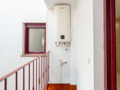 Patio interior con termo Ferroli montado en pared, suelo de baldosas terracota y barandilla metálica roja. Incluye ventana con marco burdeos y alféizar de granito, además de una puerta con marco rojo.