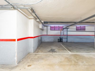 Plaza de garaje subterráneo con suelo de hormigón, paredes blancas y grises con franja roja, y tuberías vistas en el techo. Se aprecian dos pequeñas ventanas con persianas rojas.