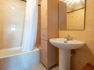 Segundo baño con lavabo de pedestal blanco, mueble alto de madera para almacenaje y bañera con cortina. Las paredes están revestidas con azulejos beige claro.