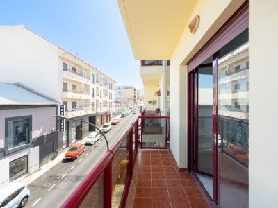 Terraza soleada con suelo de baldosas de terracota y barandilla de cristal con marcos rojos, ofreciendo vistas a una calle urbana con edificios.