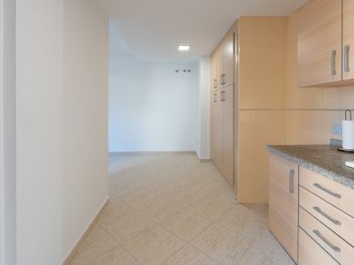 Transición desde la cocina con encimera de granito y armarios de madera clara hacia un pasillo con suelo cerámico beige y ganchos de pared.