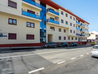 Vista exterior del edificio residencial con fachada clara, balcones de cristal azul, ventanas con persianas granates y un panel de vidrio vertical.