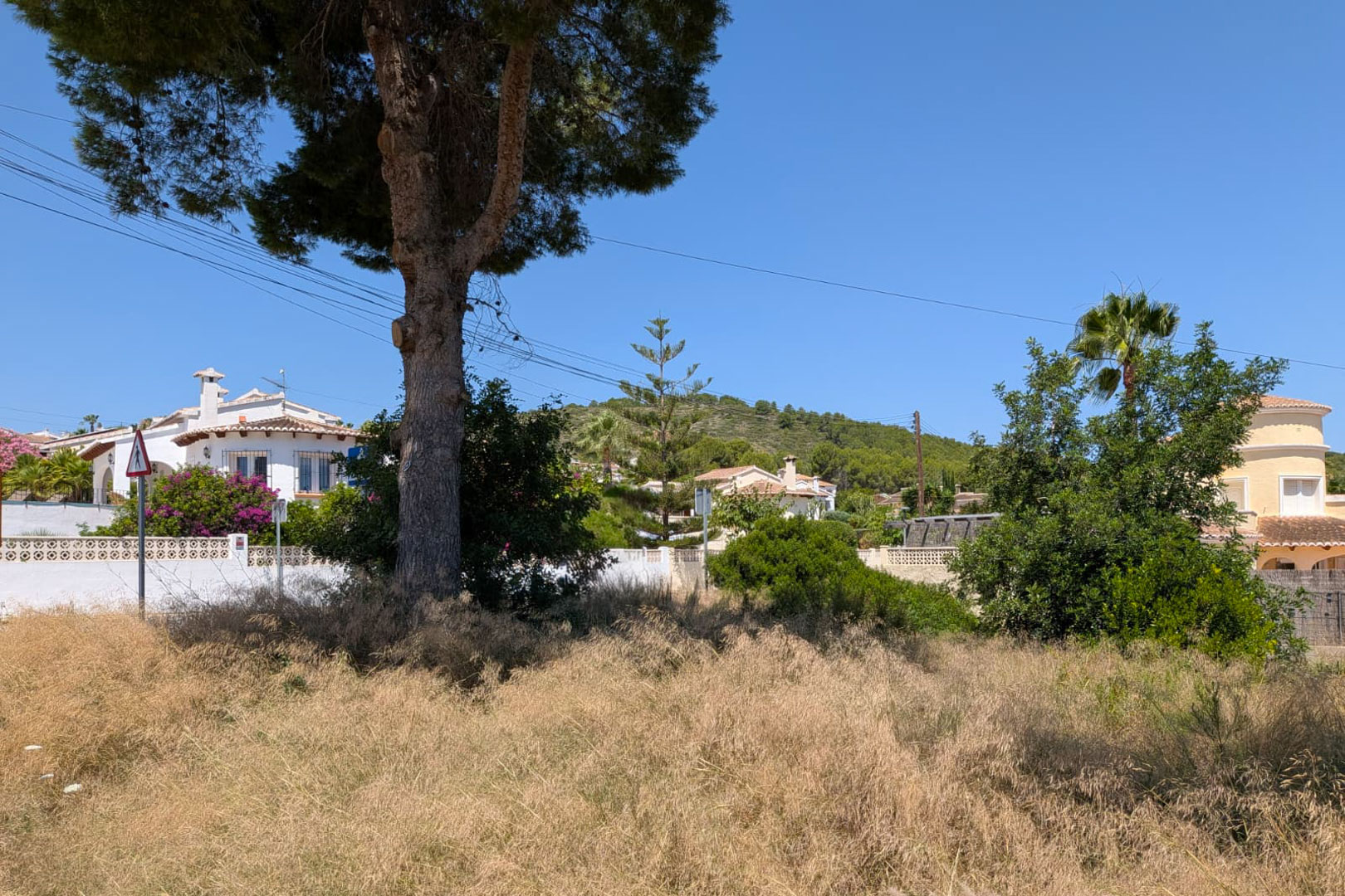 Amplia parcela con vegetación natural seca, delimitada por villas mediterráneas y una colina arbolada bajo un cielo azul despejado.