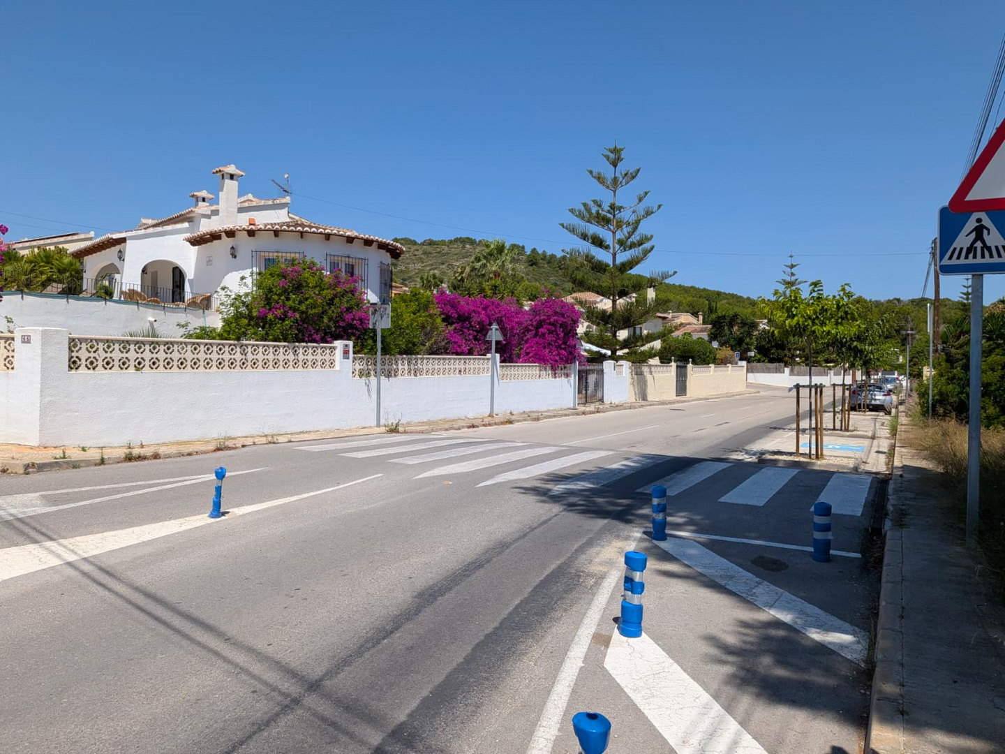 Calle residencial con paso de peatones, villa blanca tradicional y buganvillas vibrantes bajo un cielo azul claro.