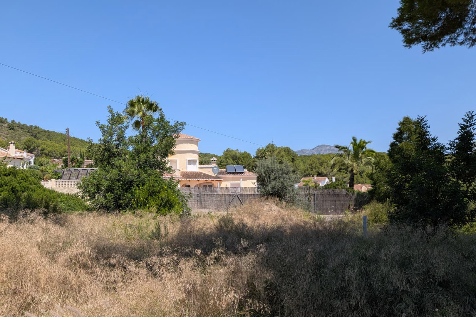 Parcela con vegetación espontánea y hierba seca, enmarcada por casas residenciales de estilo mediterráneo con tejados de teja y paneles solares, y un paisaje montañoso al fondo bajo un cielo despejado.