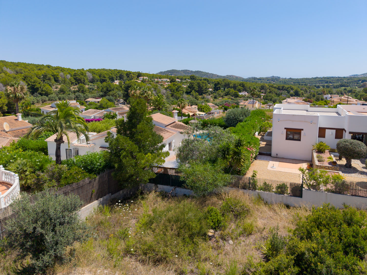 Vista aérea de la parcela sin edificar con vegetación natural, rodeada de propiedades residenciales y enmarcada por colinas verdes y cielo azul despejado.