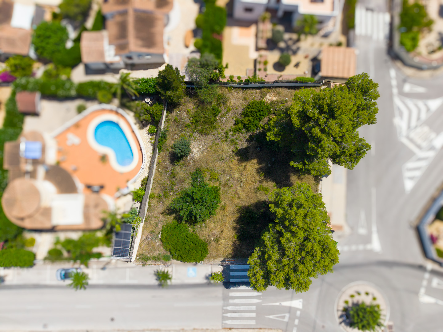 Vista aérea de una parcela edificable llana con vegetación escasa, bordeada por una calle residencial y una propiedad adyacente con piscina.