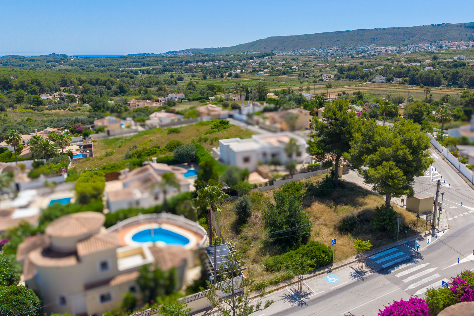 Vista aérea de una parcela residencial llana con árboles maduros, junto a una carretera con paso de cebra. Ofrece vistas lejanas al mar y a las verdes colinas circundantes.