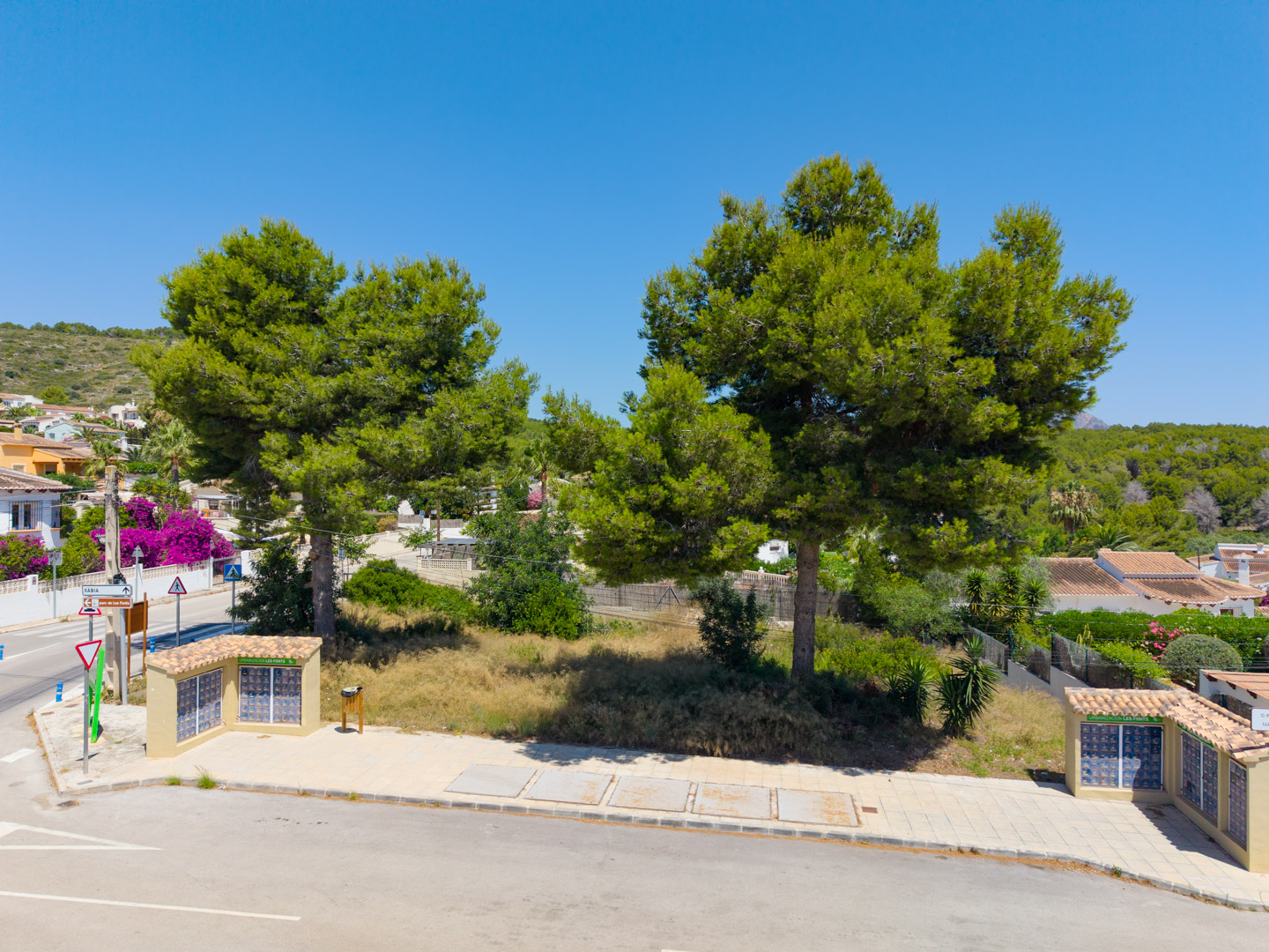 Vista aérea de una parcela soleada y sin construir en Les Fonts, con pinos maduros, calles pavimentadas y dos pequeñas estructuras con tejados de teja. Residencias y colinas verdes en el fondo.