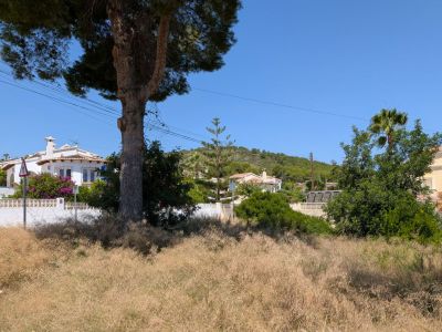 Amplia parcela con vegetación natural seca, delimitada por villas mediterráneas y una colina arbolada bajo un cielo azul despejado.
