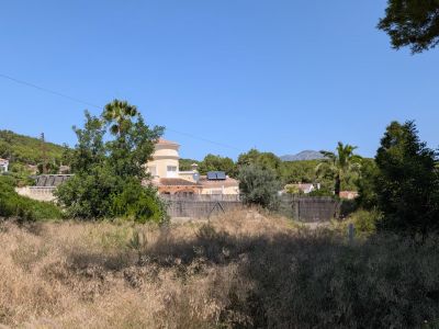 Parcela con vegetación espontánea y hierba seca, enmarcada por casas residenciales de estilo mediterráneo con tejados de teja y paneles solares, y un paisaje montañoso al fondo bajo un cielo despejado.