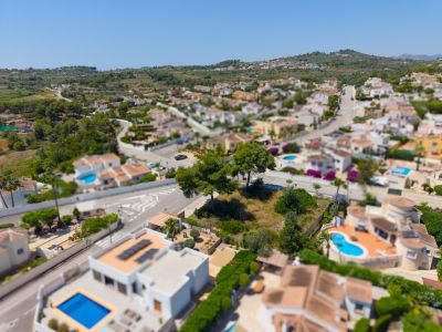 Vista aérea de una parcela llana sin edificar en urbanización residencial, delimitada por calles pavimentadas y villas con piscinas privadas, con colinas verdes al fondo.